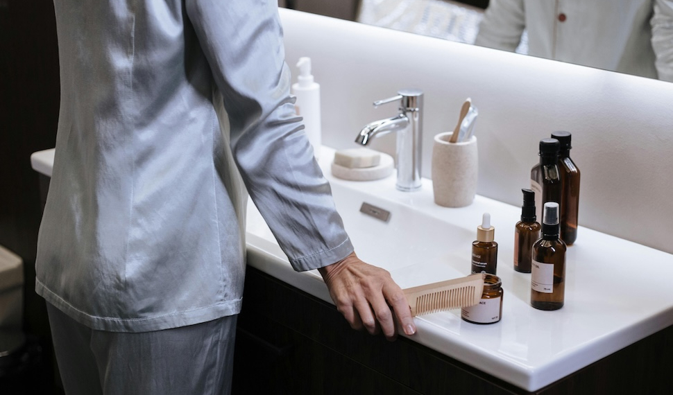 A person in pajamas stands at a modern bathroom sink, with their hand on a wooden comb. The countertop is arranged with various self-care products in amber glass bottles.